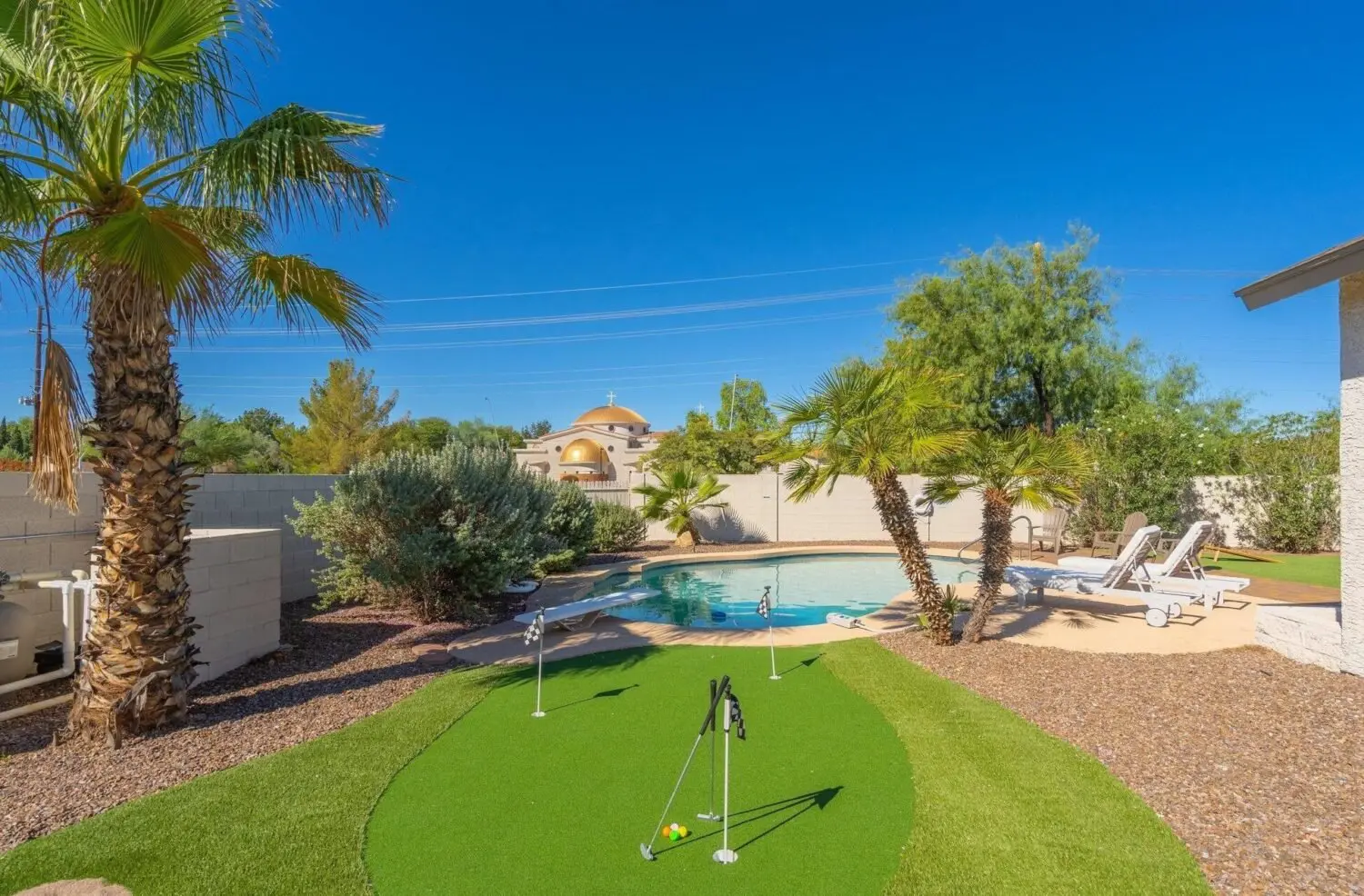 A backyard in South Mountain Village showcases a mini-golf green with three putters on artificial grass, surrounded by palm trees and desert landscaping. In the background, a swimming pool with two lounge chairs rests under a clear blue sky, promising a low-maintenance oasis.
