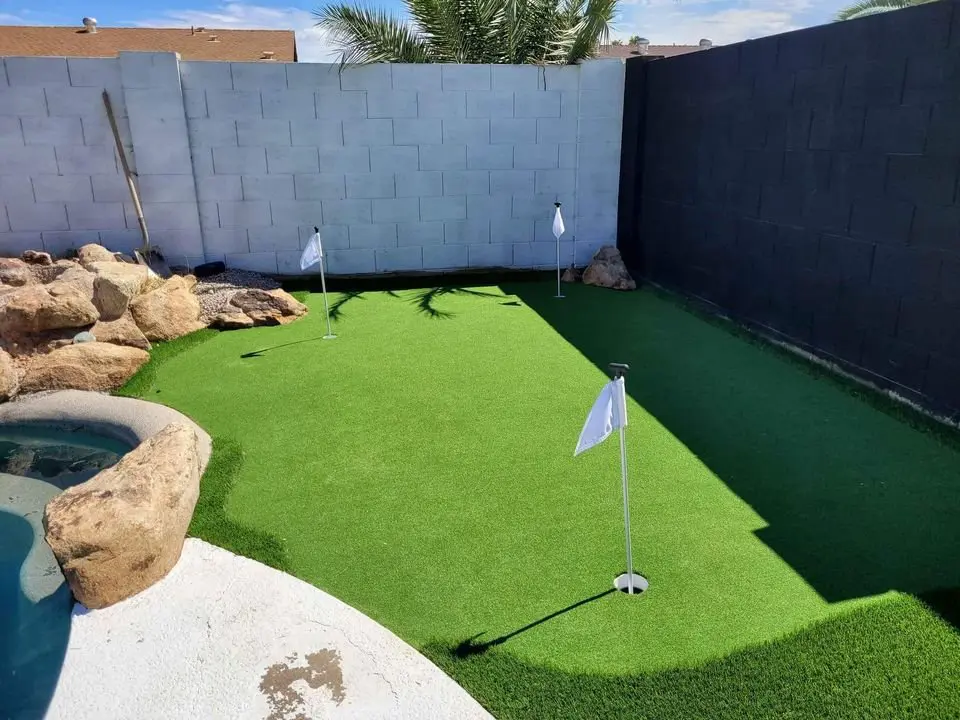 A backyard putting green crafted with premium artificial grass, featuring three small white flags on poles. It's surrounded by a stone border with rocks, and there's a partial view of a pool in the corner. A brick wall and palm fronds are under a clear sky.
