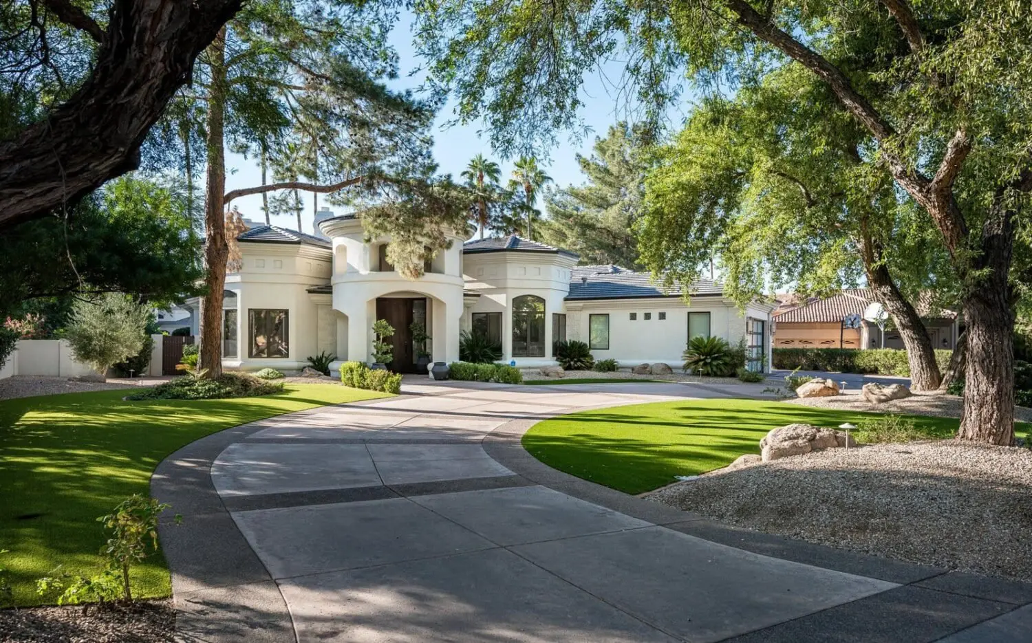 A large, elegant house with a circular driveway is surrounded by lush trees and manicured lawns featuring expertly installed artificial grass. The home boasts a prominent entrance, tall windows, and a blend of modern and classic architectural elements.