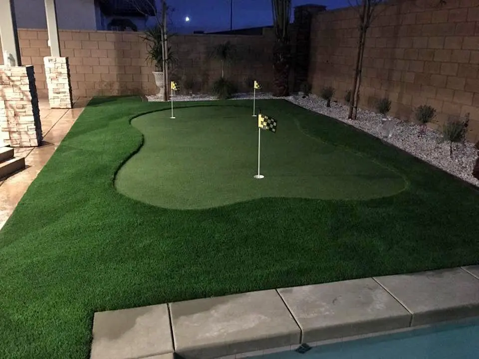A backyard paradise in Gilbert, AZ, boasts a mini golf putting green with three holes, each marked by a small flag. The green features premium artificial grass and is bordered by a stone wall on one side and a shimmering swimming pool on the other, surrounded by well-trimmed grass and decorative rocks.