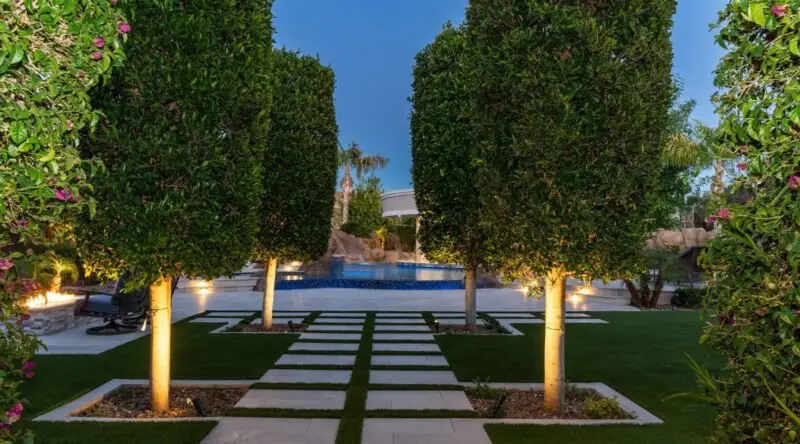 A landscaped garden path with high-quality pavers, bordered by large, neatly trimmed trees, leads to a pool in the distance. The scene is illuminated with soft lighting, enhancing the serenity of this beautifully designed outdoor space for an enchanting evening atmosphere.