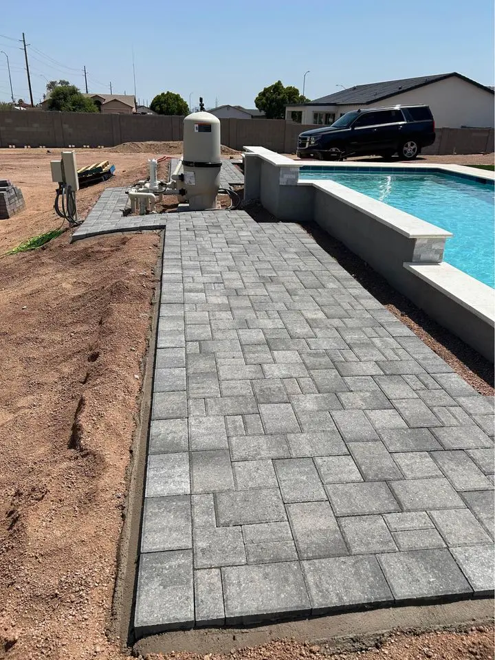 A newly constructed backyard features a swimming pool and a freshly laid gray stone walkway, showcasing high-quality pavers. A black SUV is parked nearby, with construction materials visible on the ground, hinting at ongoing outdoor space enhancements.