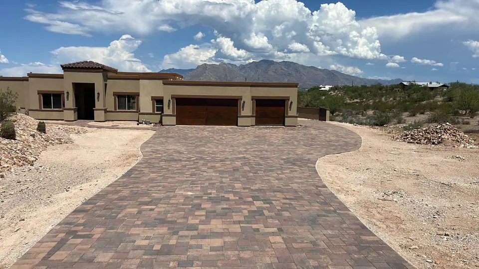 A modern house features a driveway crafted with high-quality pavers, leading to a spacious three-car garage. The dry landscape is dotted with sparse vegetation, while majestic mountains stand in the background under a partly cloudy sky.