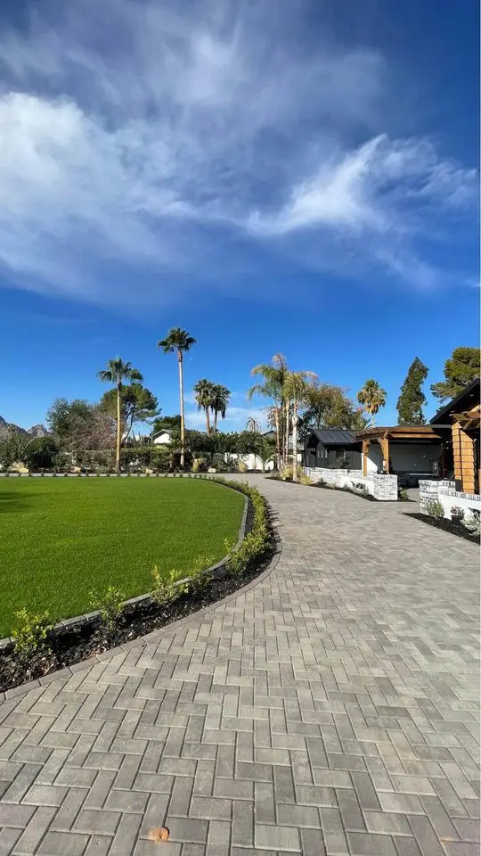 A paved walkway, crafted through expert paver installation, curves alongside a well-manicured lawn under a clear blue sky dotted with clouds. Towering palm trees accent the outdoor space, with buildings visible in the distance—a picturesque scene typical of Tempe, AZ.
