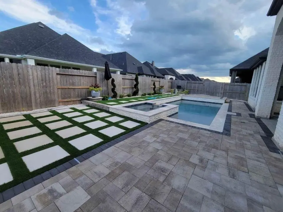 A backyard in Tempe, AZ features a modern pool with a hot tub and geometric tile design. The stylish outdoor space includes paver installation, a wooden fence against a partly cloudy sky, decorative planters, and two black umbrellas accentuating the pool area.