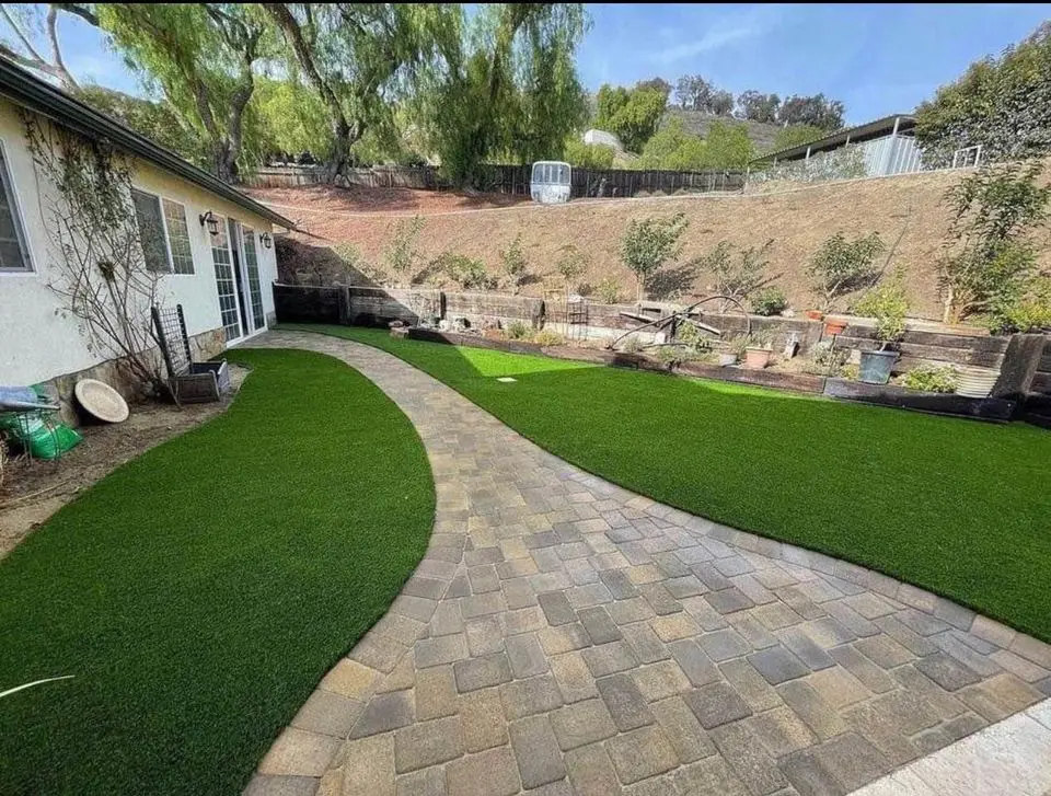 A backyard in South Mountain Village showcases a stone pathway winding through two areas of artificial grass. On the right, a garden bed rests on a hill with young trees. The left side reveals part of a house with white walls and several windows, capturing the essence of elegant landscape design.