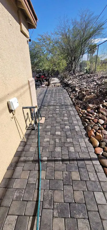 A paved walkway of high-quality pavers runs beside a beige building, lined with rocks and desert landscaping on the right. A hose stretches across the path. Trees with sparse leaves are visible against a clear blue sky, enhancing this serene outdoor space.