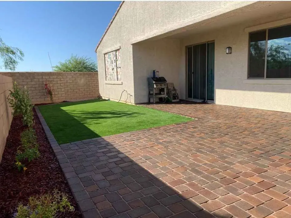 A backyard patio featuring a beautifully crafted brick surface and a small area of artificial grass, enhanced with border plants. The cozy outdoor space in Tempe, AZ includes a barbecue grill near the sliding glass door of the white house, complemented by a tall fence and clear skies in the background.