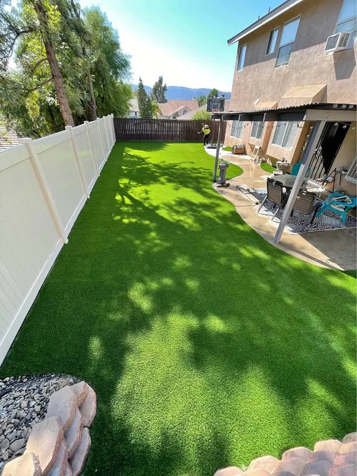 The backyard features synthetic turf enclosed by a white fence on one side and a wooden fence at the back. A patio with outdoor furniture sits to the right, while tree shadows dance on the lawn. In the foreground, a tree stands gracefully to the left.