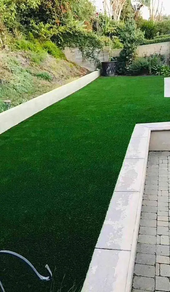 A backyard in Phoenix, Arizona, features lush green grass bordered by stone walls and a paved patio to the right. Trees and bushes line the perimeter, creating a serene outdoor space, thanks to expert artificial grass installation that ensures low maintenance.
