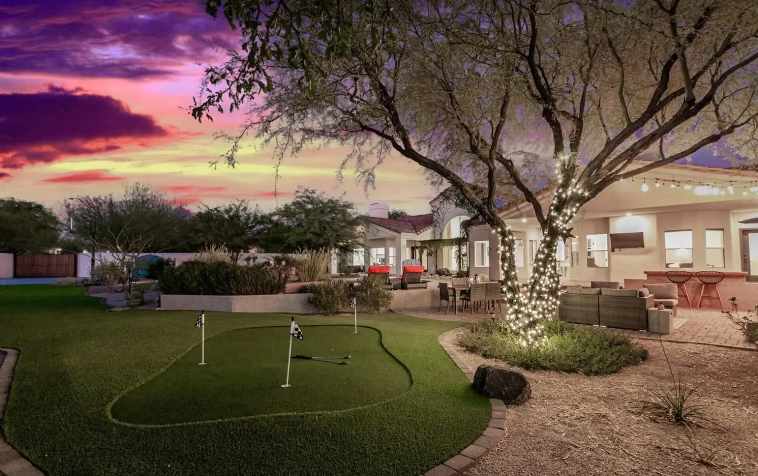 At sunset, a Phoenix, Arizona backyard glows with vibrant pink and purple hues. A small putting green of synthetic turf sits beneath a large tree wrapped in string lights, providing a serene contrast to the illuminated patio furniture and building adorned with twinkling lights.