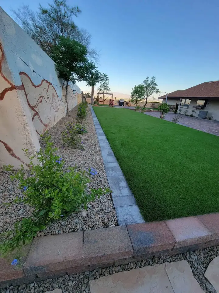 A backyard with a well-maintained putting greens lawn, bordered by a pathway and small flowering plants. A mural is painted on the left wall. The scene includes trees, a patio, and a distant view of a swing set under a clear blue sky.