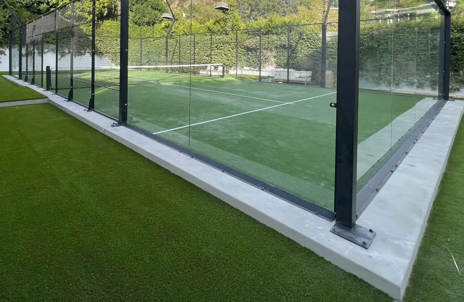 A padel court with gleaming glass walls and lush synthetic turf. Enclosed by a wire fence above the glass, this court offers benches outside, nestled amid vibrant greenery.