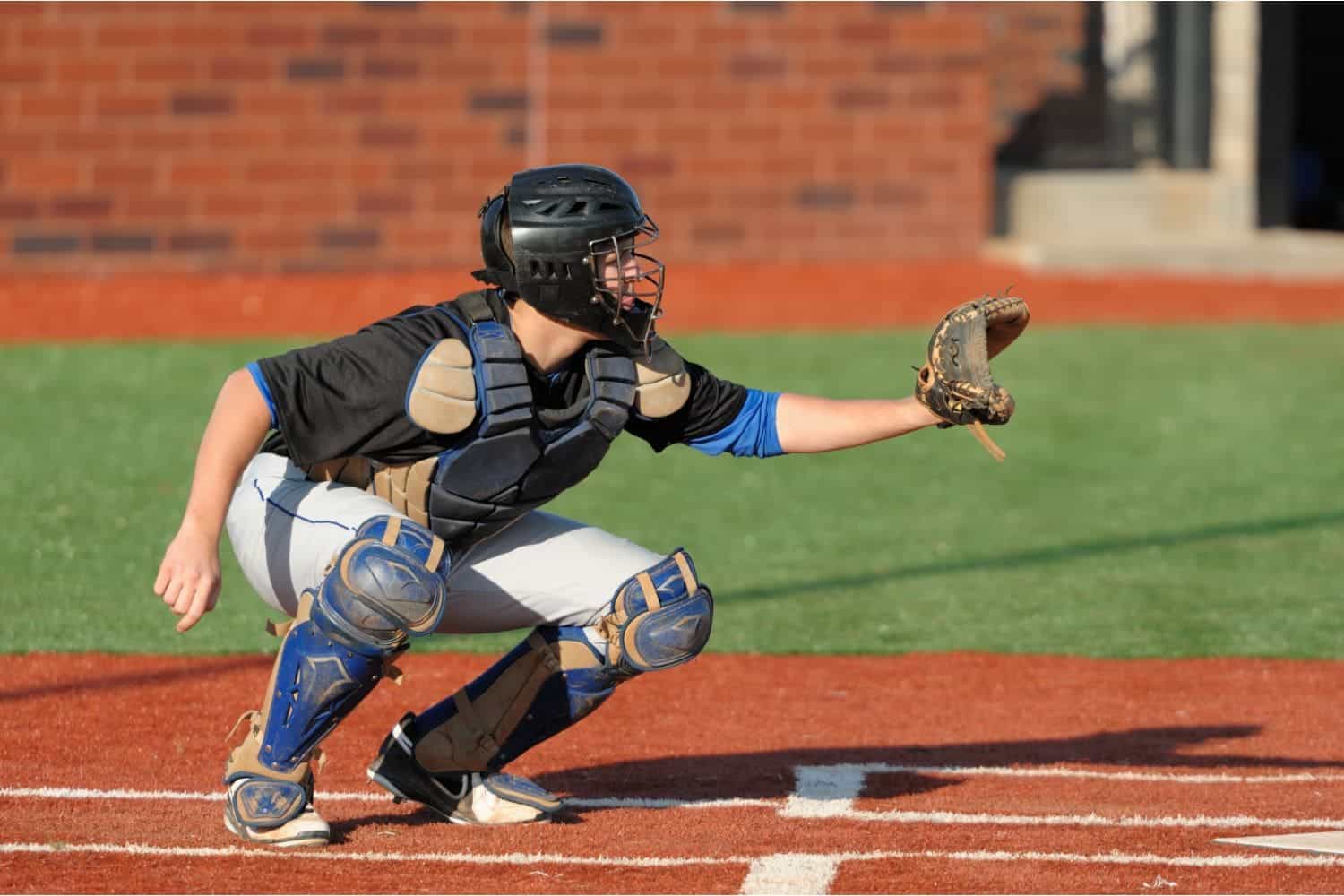 A baseball catcher crouched behind home plate wears protective gear, including a helmet, chest protector, and shin guards. They reach out with a glove to catch the ball on an immaculate artificial grass infield contrasting with the green natural outfield.