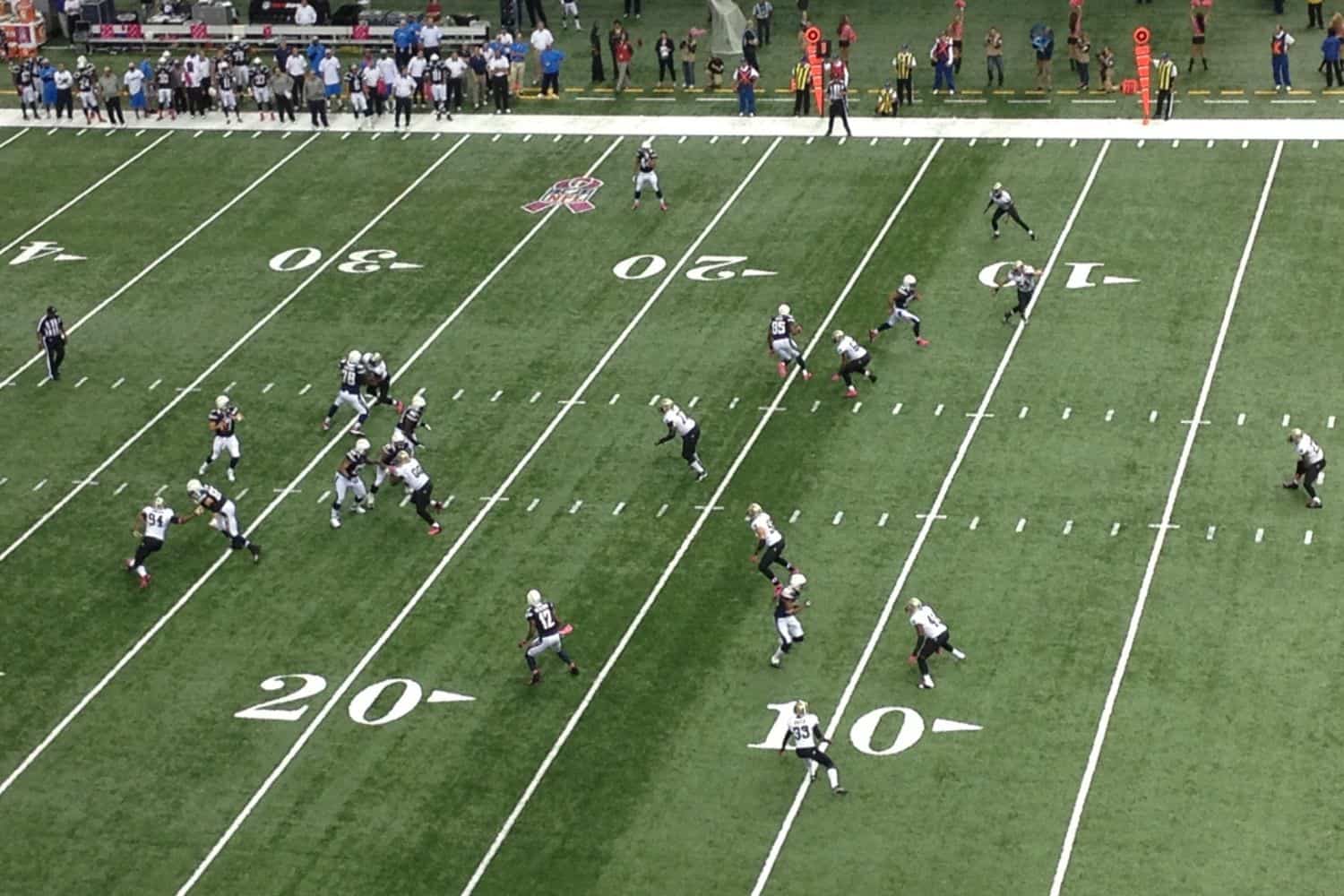 Aerial view of a football game on vibrant artificial grass, with teams in black and white uniforms. The play unfolds near the 20-yard line, players scattered across the field marked with white lines and numbers. Spectators enjoy the action from the sidelines in Gilbert AZ.